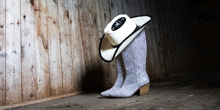 Ett par glittriga cowboyboots med en cowboyhatt ovanpå står på ett trägolv i en gammal lada.