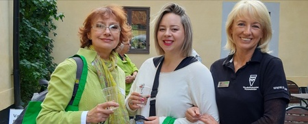 Tre kvinnor står tillsammans utomhus, två håller i plastglas och ler mot kameran.