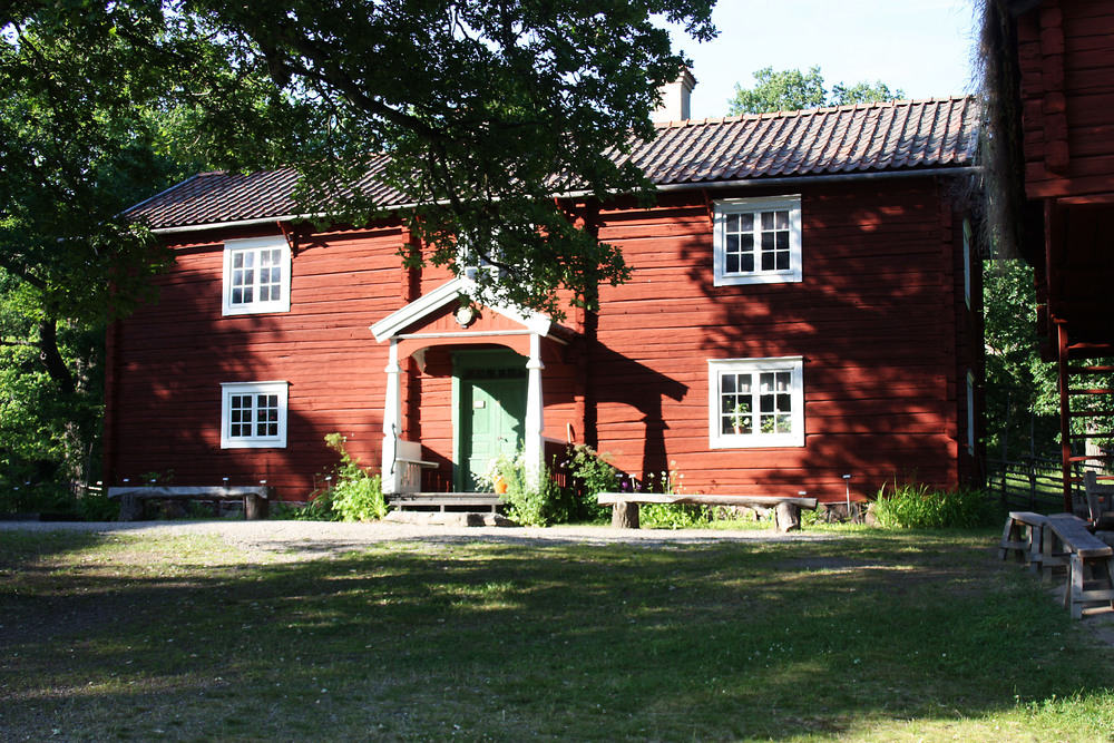 Röd trähus med vita fönster och grön dörr, omgiven av träd och gräsmatta i solsken.