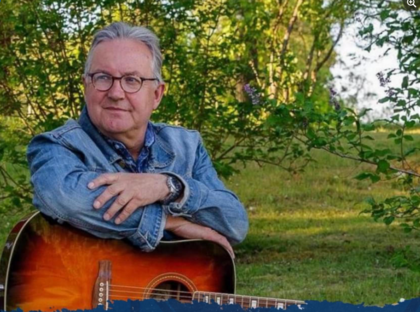 Person i jeansjacka sitter utomhus med en akustisk gitarr i knät, omgiven av grönska.