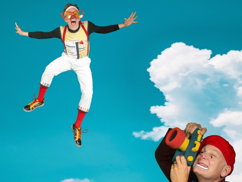 Två clowner, en skjuter upp den andra i luften med en leksakskanon mot en blå himmel med moln och höghus i bakgrunden.