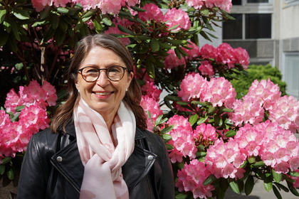 Leende kvinna med glasögon och ljus sjal står framför rosa blommande buskar utomhus.