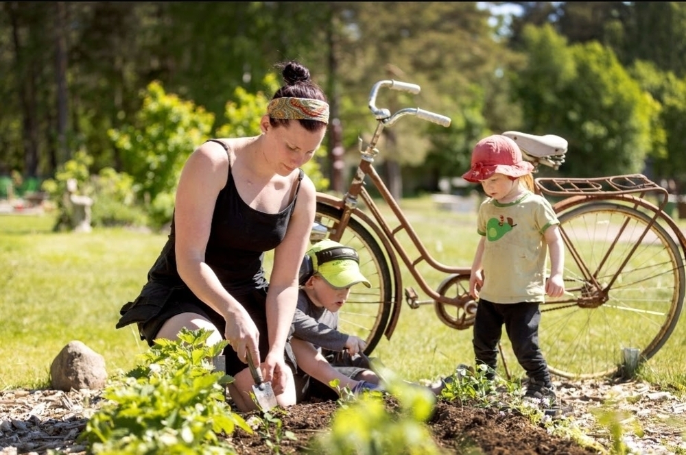 En kvinna och två barn planterar i en trädgård bredvid en cykel en solig dag.