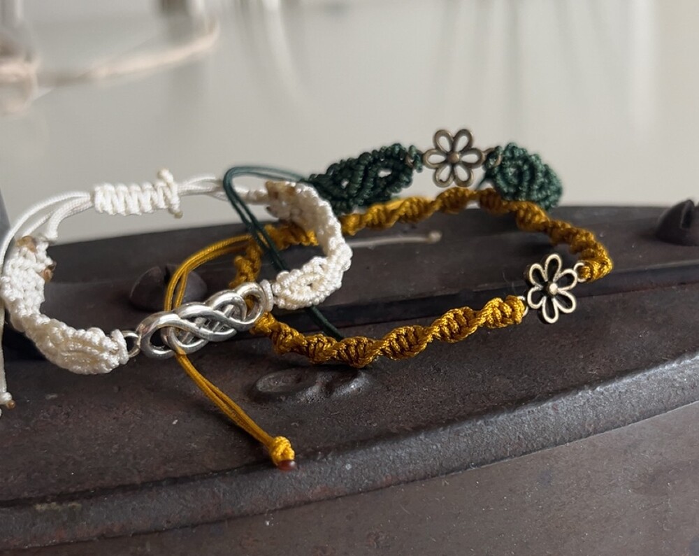 Två handgjorda armband i makramé, ett vitt och ett gult, med metallberlocker, ligger på en mörk yta.