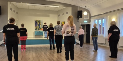 En grupp personer står i ett dansrum och följer en instruktör på en liten scen längst fram.