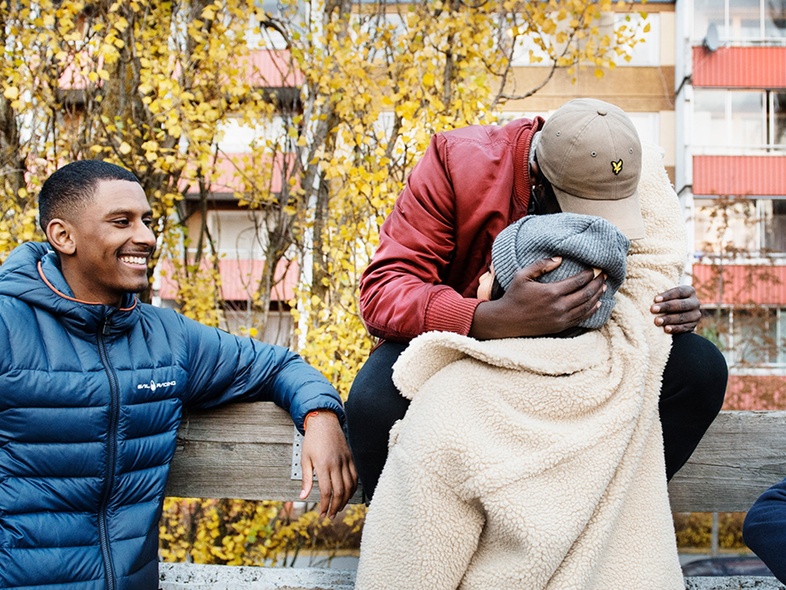 Tre personer utomhus, en sitter på ett räcke och kramar någon, en annan står bredvid och ler. Höstträd och hus i bakgrunden.