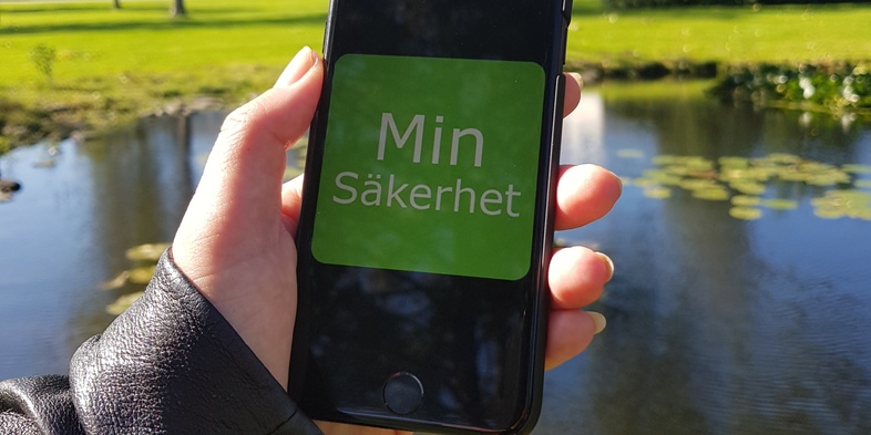 En hand håller en mobiltelefon med texten 'Min Säkerhet' framför en damm i en park.