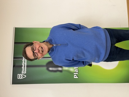 Person i blå tröja står framför en grön skylt med texten 'Studieförbundet Vuxenskolan'.