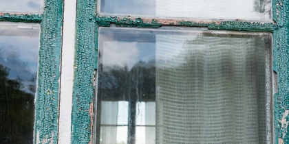 Närbild av ett gammalt fönster med avskavd grön färg och gardin på insidan.