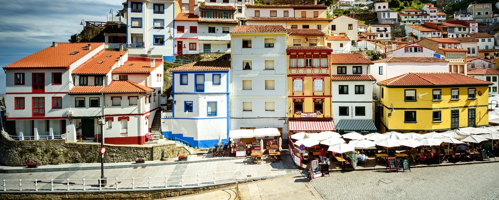 Färgglada hus i flera våningar står tätt intill varandra vid en kullerstensgata i en kuststad med grönskande backe bakom.