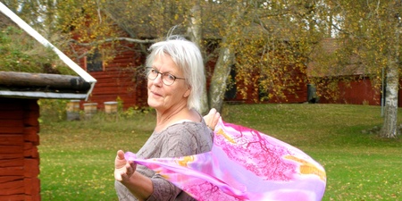 Äldre kvinna med glasögon står utomhus och håller en rosa sjal som blåser i vinden.