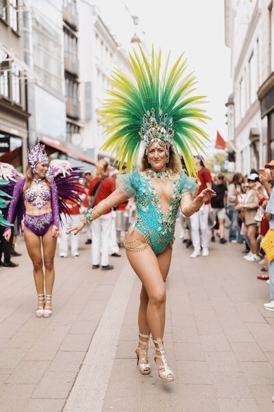 Kvinna i färgglad karnevalsdräkt med fjädrar dansar på en stadsgata under en parad.