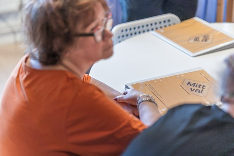 En person med glasögon sitter vid ett vitt bord. På bordet ligger pärmar med Mitt val-logotyp.