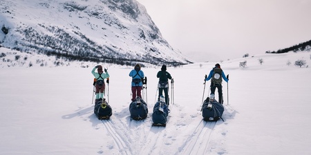 Fyra personer med pulkor står på skidor i ett snötäckt fjällandskap med berg i bakgrunden.