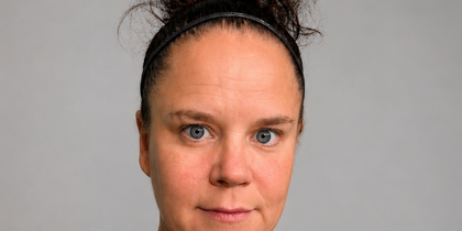 Kvinna med mörkt lockigt hår i tofs och svart pannband, tittar rakt mot kameran mot grå bakgrund.