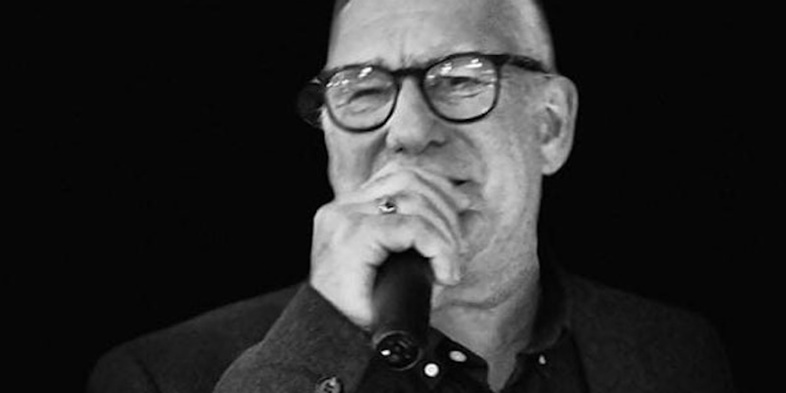 Man med glasögon och kavaj håller en mikrofon och talar, svartvit bild.