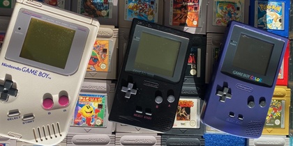 Tre Game Boy-konsoler och flera spelkassetter ligger på en blå matta i solljus.