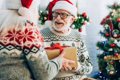 Två äldre personer i jultröjor och tomteluvor utbyter en inslagen present framför en julgran.