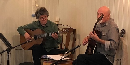 Två personer spelar akustisk gitarr på en liten scen i ett mysigt rum med vita träväggar.