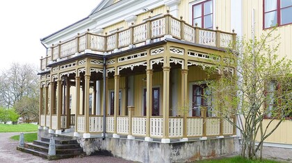 Gul trävilla med snickarglädje och stor veranda, omgiven av grönska.