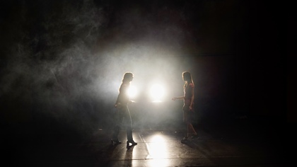 Två personer står mittemot varandra på en mörk scen med starkt motljus och rök.