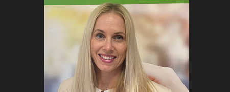 Leende kvinna med långt blont hår framför en suddig bakgrund och en grön skylt ovanför.