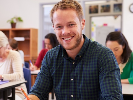 Leende man sitter vid skolbänk i klassrum, skriver i anteckningsbok. Andra vuxna studenter i bakgrunden.