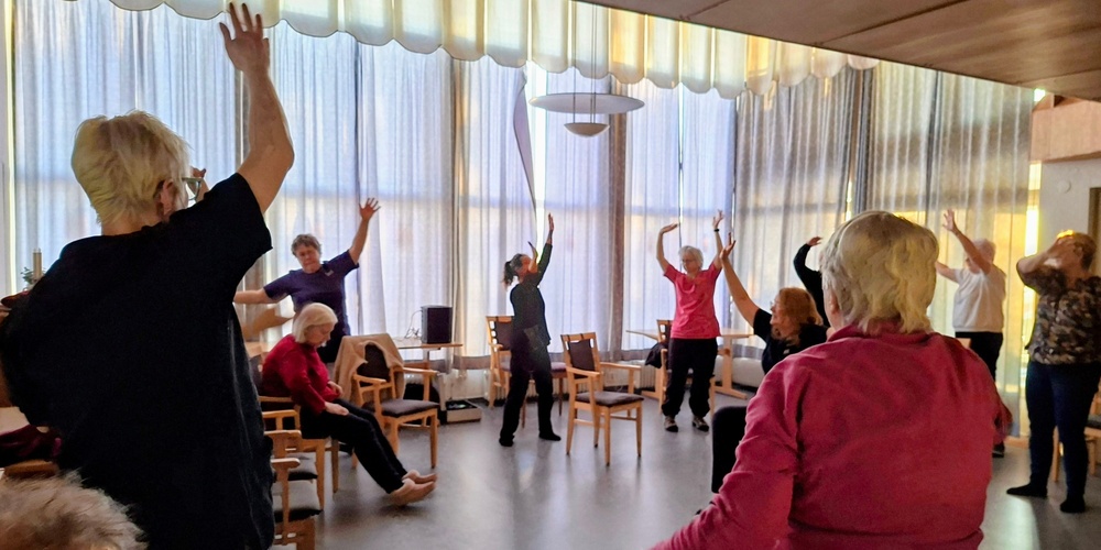 En grupp äldre personer deltar i sittande och stående gymnastik i ett ljust rum med stora fönster.