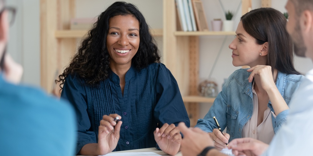 Fyra personer sitter runt ett bord och har ett möte, en kvinna i fokus ler mot kameran.