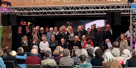 Kör och musiker uppträder på scen inför sittande publik i en inomhuslokal med dekorerad vägg.