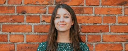 Ung kvinna med långt brunt hår står framför en tegelvägg och ler mot kameran.