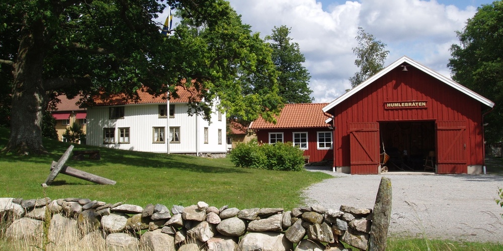 Gårdsmiljö med röd lada, vit byggnad, stengärdsgård och grönskande träd under blå himmel.