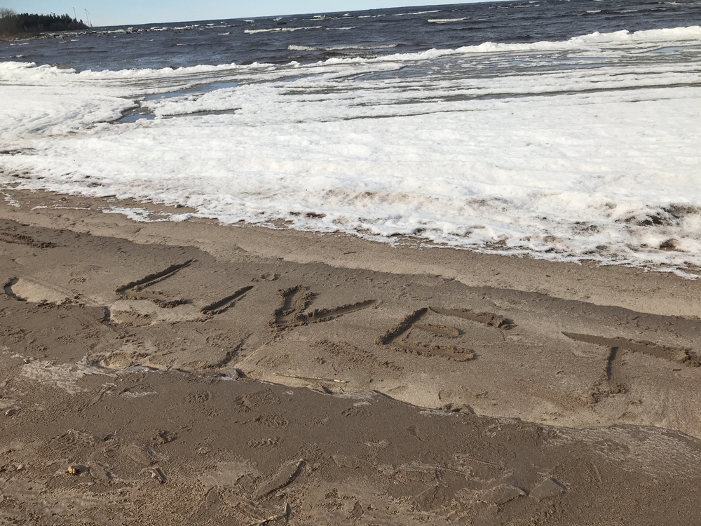 Texten 'LIVET' är skriven i sanden på en strand med vågor och skum i bakgrunden.