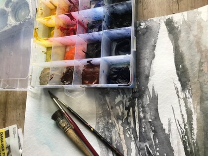 Akvarellfärger, penslar och ett påbörjat konstverk på ett bord.