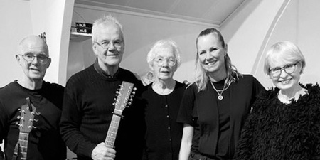 Fem personer står tillsammans inomhus, två håller akustiska gitarrer, alla är klädda i mörka kläder.