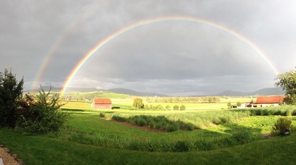 Landskap med gröna fält, bondgårdar och en tydlig dubbel regnbåge mot en molnig himmel.