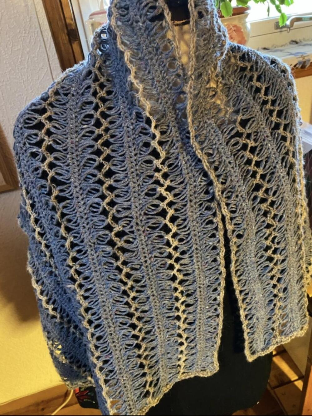 En blå, handstickad sjal med luftiga öglor visas på en provdocka inomhus.