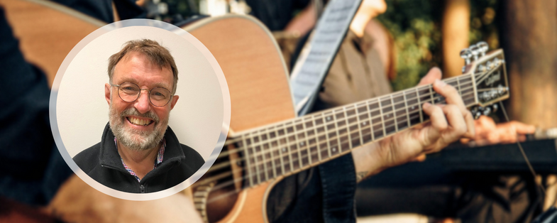 En leende man med glasögon i en cirkel framför en bakgrund av någon som spelar akustisk gitarr utomhus.