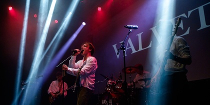 Ett band uppträder på scen med färgglada strålkastare och instrument, texten 'VALVET' syns i bakgrunden.