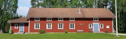 Rött trähus med vita knutar och blå dörrar, omgiven av gräsmatta och träd.