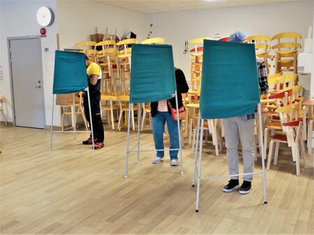 Tre personer röstar i valbås i ett rum med staplade stolar i bakgrunden.