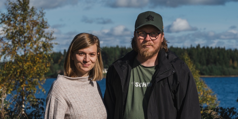 En kvinna och en man står utomhus vid en sjö med skog i bakgrunden under en solig dag.