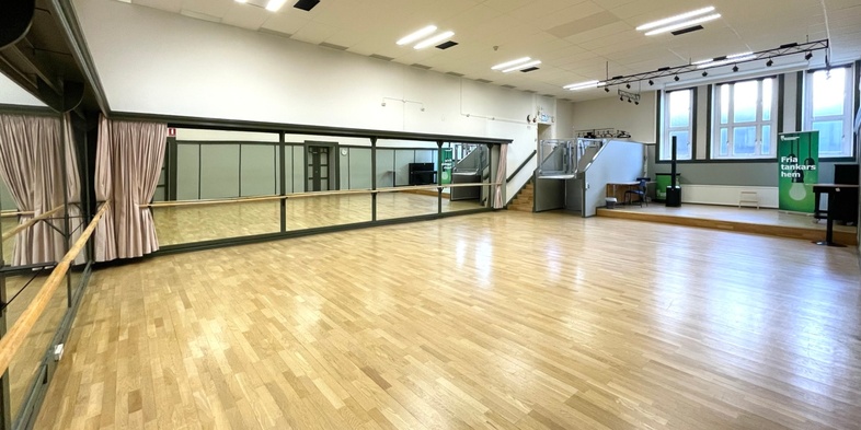 Ljust dans- eller träningsrum med trägolv, stora speglar och scen i ena änden.
