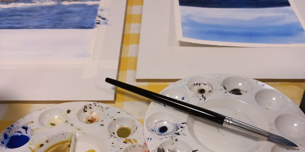 Två målarpaletter med färg, pensel, tuber med akrylfärg och två målningar på ett bord med gulrutig duk.