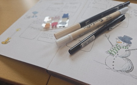 Anteckningsbok med teckning av en snögubbe, pennor och färgglada klistermärken på ett skrivbord.