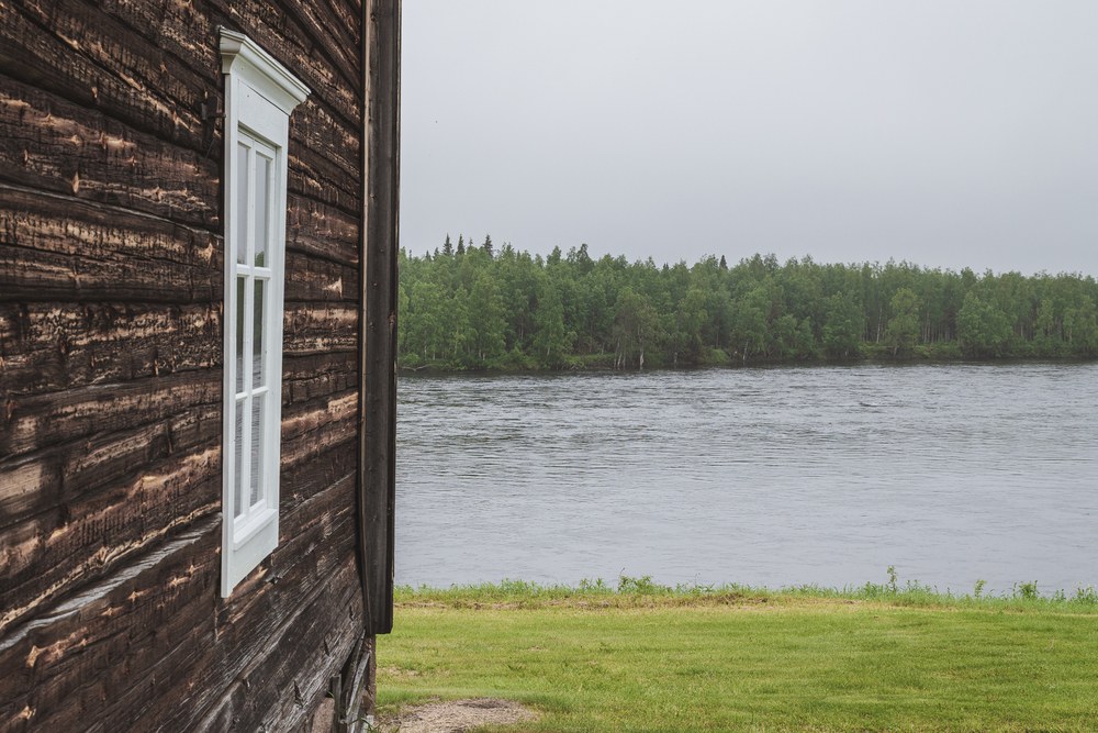 En trävägg med vitt fönster syns till vänster, med en flod och skog i bakgrunden under en mulen himmel.
