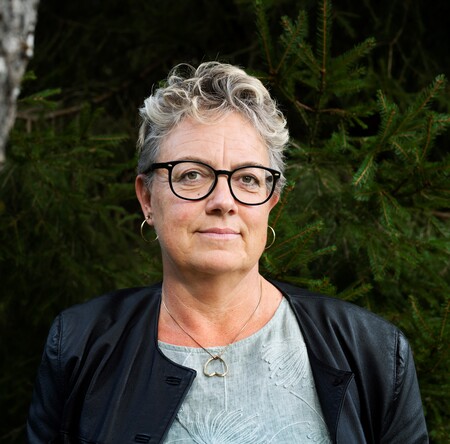 Kvinna med kort, lockigt grått hår och glasögon står utomhus framför gröna träd, bär jacka och ljus topp.