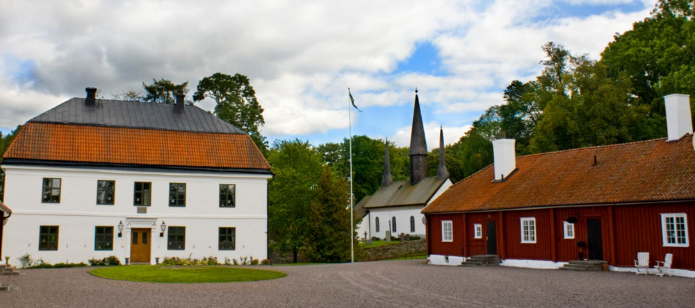 Gårdsmiljö med vit herrgårdsbyggnad, röd stuga och kyrka med spira omgivna av grönska.