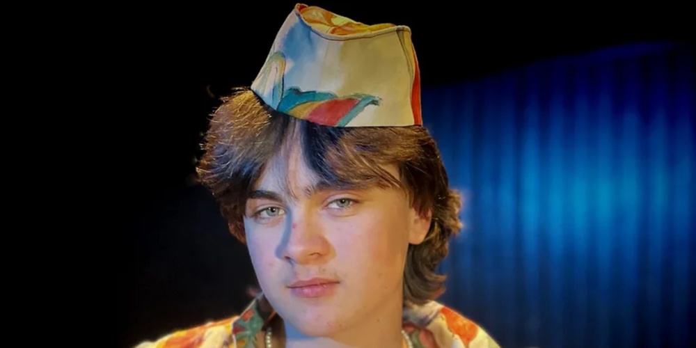 Ung person med färgglad skjorta och hatt poserar mot mörk bakgrund.