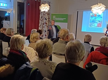 En grupp äldre personer sitter i ett rum och lyssnar på en presentation med bildspel.